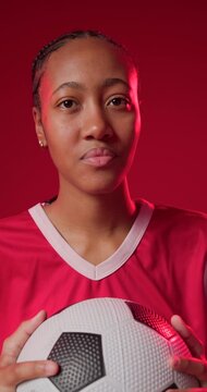 As camera rolling African American woman in red jersey holding soccer ball, shifting gaze in studio