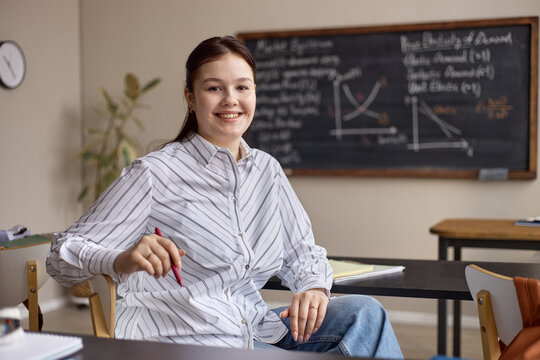 Portrait of Caucasian young adult woman sitting in classroom, holding pen, smiling at camera with math board behind. Suitable for education marketing, tutoring services, school promotion
