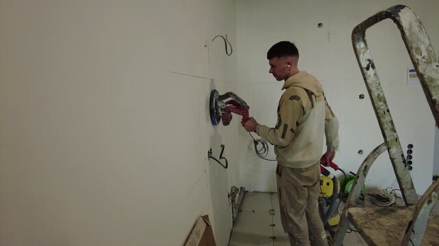 A man is working on a construction site. He's using a power tool to strip a wall. He's wearing headphones and is focused on his work. Construction materials and equipment are visible in the background
