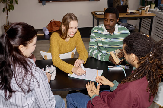 Multiethnic young adults collaborating around table, reviewing notes and discussing ideas in study group. Useful for education marketing, teamwork, tutoring, campus life campaigns