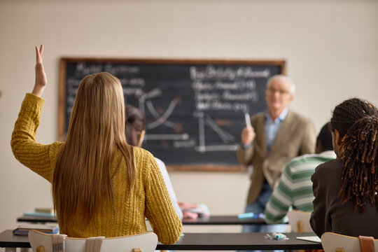Teenage girl raising hand during classroom lesson while senior male teacher explaining graphs on chalkboard, showing active learning and student engagement. Useful for education marketing