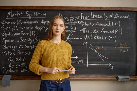 Portrait of young adult Caucasian woman standing before chalkboard, teaching economics concepts and market equilibrium. Ideal for education marketing, tutoring services, academic content