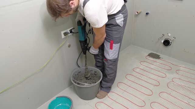 Construction worker preparing tile surface, mixing mortar substrate for flooring, underfloor heating mat. Workman combines adhesive in renovation setting. Operating power tool during bathroom upgrade
