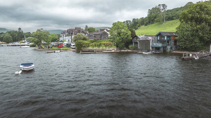 Fototapeta premium Boats and buildings line a scenic lake shore on a cloudy day, Lake District, UK