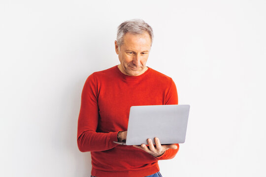 A man stands against a white wall while he uses a laptop. He is wearing a red sweater and appears focused on his work. The setting is simple with no distractions.
