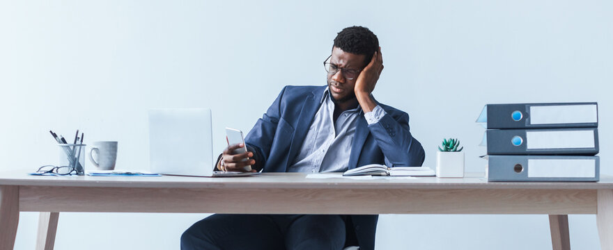A young man is sitting at his desk in an office looking worried while holding his smartphone. He appears to be experiencing stress or frustration. The workspace has a laptop, papers, and a plant.