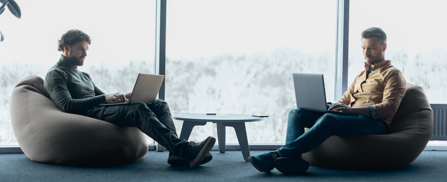 Two men sit in bean bag chairs while working on laptops. They are focused on their screens. A large window shows a view of trees outside. Natural light fills the room.