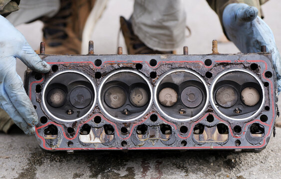 Disassembled cylinder head with combustion chambers facing up, showing valves and gasket area, with gloved hands pointing to sealing issues and wear. 2026