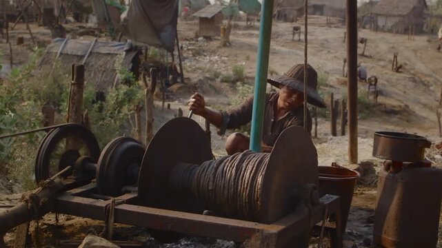 Burmese man operating a rusty manual winch to extract crude oil in a traditional oil field