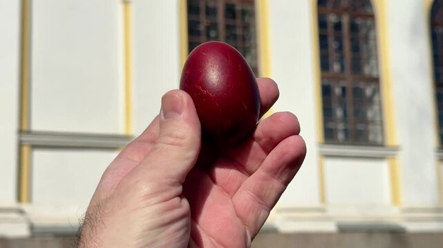 Hand holding a red Easter egg with a blurred church building in the background. Concept of Easter celebration, spring and traditional symbolism