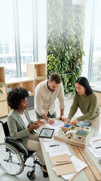Diverse team of architects and engineers, including a woman in a wheelchair, collaborating on a sustainable building model with solar panels in a modern, eco-friendly office environment