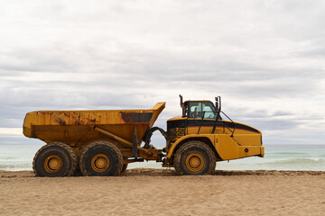 Obraz premium Large yellow dump truck standing on sandy beach near the sea under cloudy sky. Heavy machinery used for coastal work.