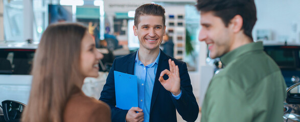 A salesperson talks with a couple inside a car dealership. The salesperson smiles and gestures positively, while the couple listens. Cars are visible in the background.