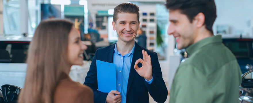 A salesperson talks with a couple inside a car dealership. The salesperson smiles and gestures positively, while the couple listens. Cars are visible in the background.