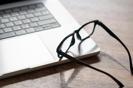 Silver laptop keyboard sits on a wooden table. Black eyeglasses lean against the laptop's trackpad. Natural light creates a serene workspace mood. This setup implies concentration during work or study