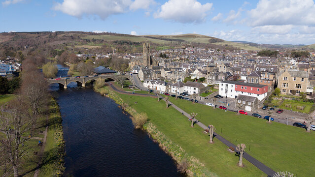 A picturesque perspective looking over the heart of Peebles, showing the town's connection to the River Tweed. The water curves gracefully past the historic stone buildings and lush riverside parks, i