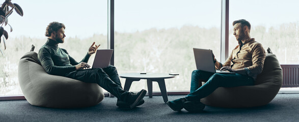 Two men sit facing each other in an office space with large windows. They are working on laptops and having a discussion. The setting has a modern design with natural light coming in.