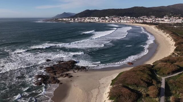 Aerial view of the beach where dark blue ocean meets the sandy shore, with the town nestled against the green hills, Vila Praia de Ancora, Portugal.