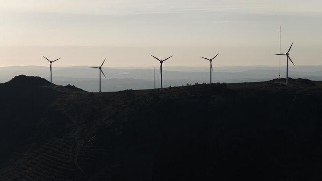 Aerial view of several wind turbines standing tall on a dark hill, harnessing wind power against a backdrop of distant mountains, Amarante, Portugal.