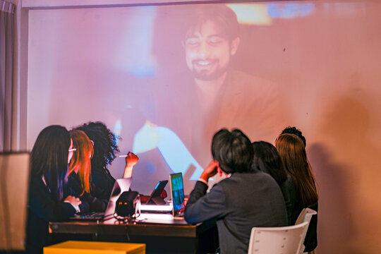Professional IT squad analyzing machine learning data with a remote expert via projector. Modern tech workplace featuring cybersecurity dashboards and global threat sharing discussions.