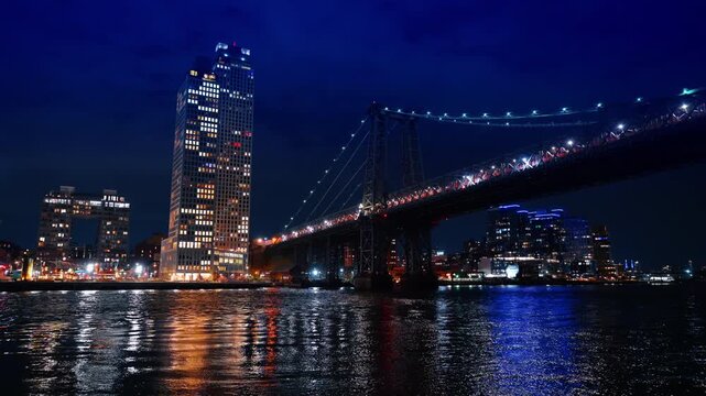 City lights reflect in the river at night. Low angle view at the Williamsburg Bridge from the waterscape.