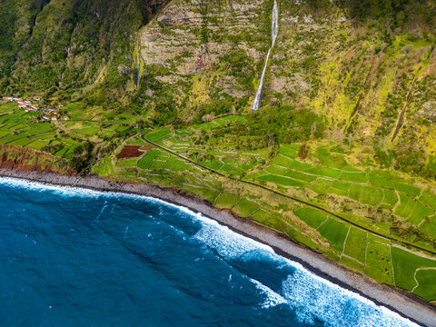 Aerial view of a dramatic coastline where the rugged, verdant cliffs meet the deep blue sea, with a waterfall cascading down the slopes, Faja Grande, Azores, Portugal.