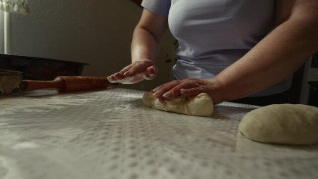 Caucasian woman in light blue shirt kneads dough on a countertop, using hands and a rolling pin, with a kitchen setting and baking tools visible in the background