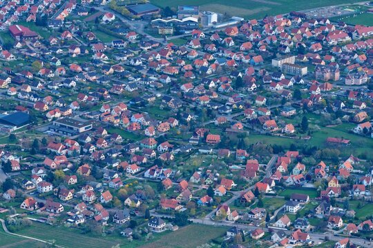 Aerial view of the dense tapestry of red-tiled roofs contrasting with lush green spaces, a sprawling suburban landscape under a soft, diffused light, Kintzheim, Grand Est, France.