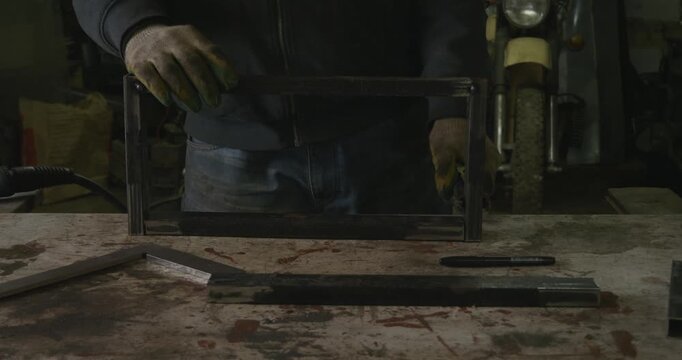 Skilled craftsman wearing protective gloves meticulously assembling a rectangular metal frame on a workbench, preparing the steel profile pipes for welding in a dimly lit industrial garage.