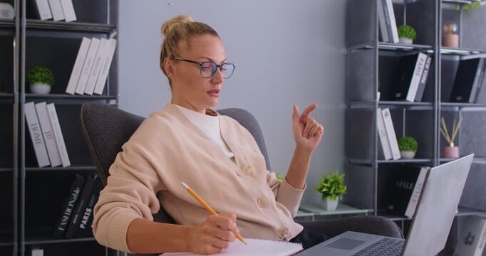 Focused woman handles online meeting via laptop in office, managing business communication, engaging in internet video call, enhancing work efficiency with professional interaction and technology use.