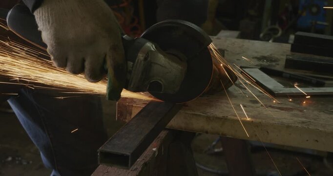 Professional worker in protective gloves cutting a rectangular steel pipe with an angle grinder, creating a shower of hot sparks flying in the industrial workshop during metalworking.