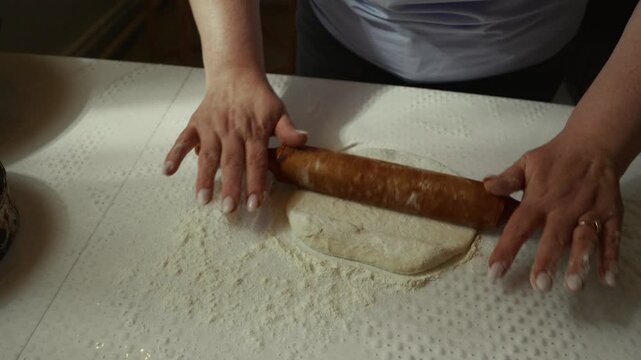 Woman prepares dough by rolling it out with a wooden rolling pin on a floured surface in a well-lit kitchen, showcasing the process of making fresh bread