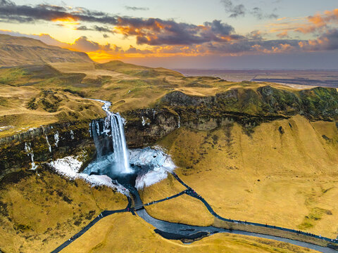 Aerial view of cascading waterfall plunges into icy pool amidst rugged landscape, golden fields contrasting with dark cliffs, Seljalandsfoss, Rangarthing eystra, Iceland.