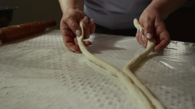 Female baker skillfully twists and shapes dough into a braided form on a textured surface, with a rolling pin and bowl visible in the background