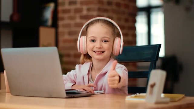 A girl with light skin sits at a table in a bright room. She is using a laptop with headphones on. She gives a thumbs-up and smiles. The scene shows a comfortable home learning environment.
