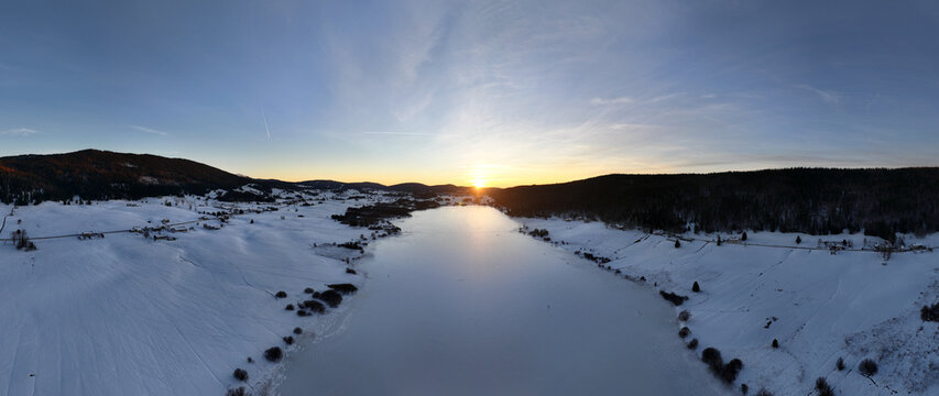 Aerial view of a frozen lake reflecting the vibrant sunset, bordered by snow-covered landscapes and dark forests, Les Rousses, Bourgogne-Franche-Comte, France.