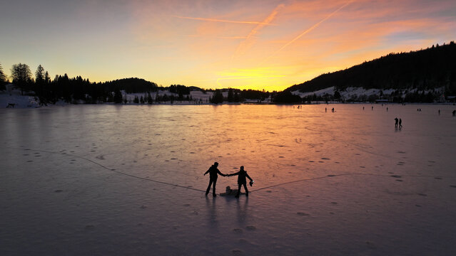 Aerial view of figures skating on the frozen lake with a backdrop of silhouettes of trees against the colorful sunset sky, Les Rousses, Bourgogne-Franche-Comte, France.