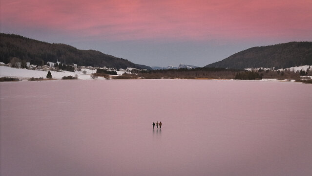 Aerial view of three figures stand silhouetted on the vast, frozen expanse of the lake under the soft, pink hues of the winter sky, Les Rousses, Bourgogne-Franche-Comte, France.