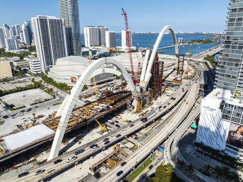 Aerial view of the construction of the dramatic arch of the signature bridge over I-95, a symphony of steel and concrete amidst the vibrant cityscape and bay waters, Miami, Florida, USA.