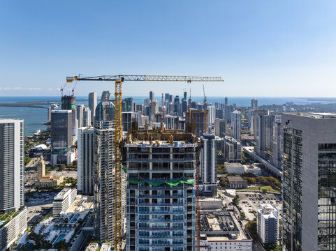 Aerial view of a skyscraper under construction piercing the skyline, with cranes looming over the cityscape, against the backdrop of Biscayne Bay, Miami, Florida, USA.