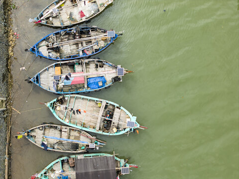 Aerial view of weathered fishing boats, their teal and blue paint peeling under the harsh sun, resting beside a muddy shore, Jepara, Central Java, Indonesia.