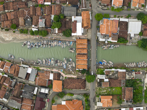 Aerial view of boats docked along the river, juxtaposed against the rustic red rooftops of the buildings, creating a vibrant tapestry of urban life, Jepara, Central Java, Indonesia.
