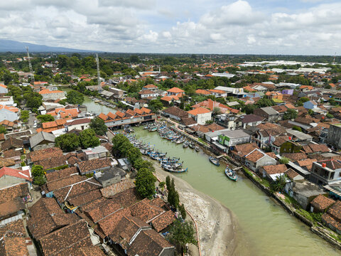 Aerial view of boats clustered at the floating fish market, a vibrant scene of commerce amidst the red-tiled rooftops and lush greenery, Jepara, Central Java, Indonesia.