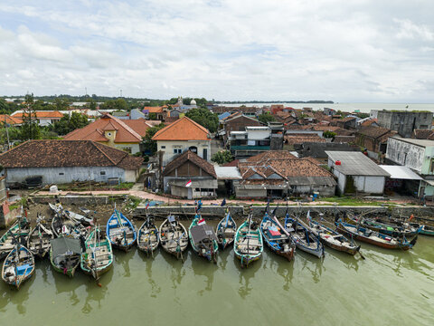 Aerial view of colorful boats clustered along the Jepara Floating Fish Market, contrasting with the earthy tones of the buildings and the sea, Jepara, Central Java, Indonesia.