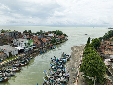 Aerial view of boats docked along the riverbanks, with the town's red-tiled roofs contrasting against the cloudy sky, Jepara, Central Java, Indonesia.
