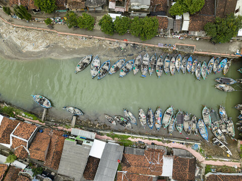 Aerial view of boats docked along the river contrasting with the red-tiled rooftops of buildings, casting shadows in the afternoon sun, Jepara, Central Java, Indonesia.