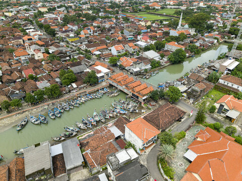 Aerial view of fishing boats docked along the murky riverbanks beneath the bridge, contrasting with the surrounding red-tiled roofs, Jepara, Central Java, Indonesia.
