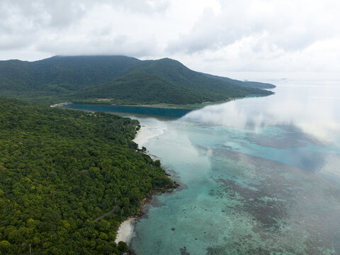 Aerial view of the lush green coastline meeting the turquoise waters under a cloudy sky, reflecting the horizon, Karimunjawa, Central Java, Indonesia.