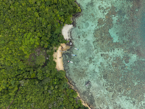 Aerial view of lush green forests meeting the turquoise sea at Karina Beach, revealing boats nestled in the sandy shore, Karimunjawa, Central Java, Indonesia.