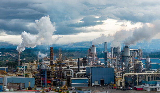 Aerial view of industrial complex billowing smoke into the cloudy sky, a stark contrast against the distant hills, Medellin, Colombia.